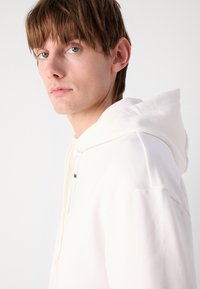 Biely hoodie s šnúrkou, ktorý má mäkkú textúru a voľný strih. Minimalistický dizajn bez viditeľných log a vzorov.