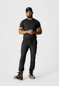 Schwarzes T-Shirt und schwarze Jogginghosen, kombiniert mit braunen Stiefeln. Die Mütze ist schwarz mit einem grünen Akzent und einem weißen Logo.