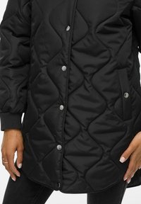 Μαύρο επενδυμένο μπουφάν με κυματιστό σχέδιο ραφής, κουμπιά με κλίπ μπροστά και χέρια που στηρίζονται κοντά στις τσέπες του γοφού.