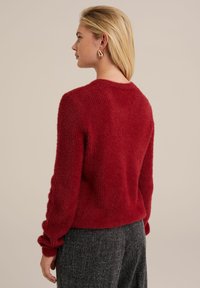 Sarkana, pūkaina adīta džemperis ar apaļu kaklu un garām piedurknēm; ir ribota tekstūra un nedaudz pārmērīgi plaša piegriezuma. Valkāts ar pelēkiem biksēm.