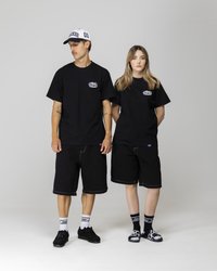 T-shirt in cotone nero con logo, pantaloncini di jeans neri con cuciture a contrasto, abbinati a scarpe nere e calze a fantasia. Cappelli indossati da entrambi.
