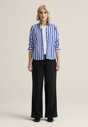 Kvinde stående med afslappede hænder, iført blå- og hvidstribet skjorte over hvid top, sorte bukser med vide ben og sorte sko.