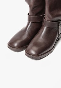 Botas de cuero marrón oscuro hasta la mitad de la pantorrilla, con punta cuadrada redondeada, detalle de correa y hebilla, y suelas gruesas y texturizadas sobre un fondo blanco.