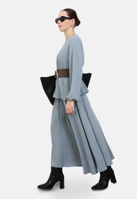 Langes, hellblaues Kleid im Peplum-Stil, brauner Taillengürtel, schwarze Handtasche und schwarze Stiefeletten. Als Accessoires werden runde schwarze Sonnenbrillen getragen.