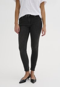 Jeans negros ajustados de mezclilla, con una cintura de altura media, cinco bolsillos y detalles de costura sutiles. Combinados con zapatos negros de punta afilada.