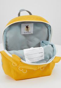 Sac à dos en tissu jaune avec un intérieur bleu, comprenant une poignée supérieure, une étiquette, et des détails de couture blanche sur l'extérieur.