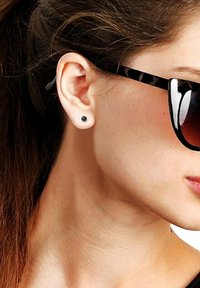 Boucle d'oreille noire en acier, contrastant avec le teint de la peau. Cheveux coiffés, et lunettes de soleil avec un design noir et écaille de tortue.