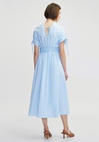 Robe bleu clair, longueur genou, avec des manches courtes froncées et une taille smockée. Présente un dos en V avec fermeture à nouer et une jupe fluide.