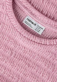 Tessuto rosa testurizzato di una maglietta per bambini con design arricciato e scollo a costine, con etichetta di cura visibile.