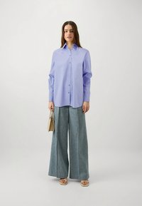 Light purple button-up shirt with long sleeves, paired with wide-leg denim trousers. Model holds a small beige handbag. Simple, clean design.