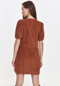 Frau in einem braunen Cordkleid mit Puffärmeln, steht mit dem Rücken zur Kamera und zeigt den Reißverschluss an der Rückseite vor einem schlichten weißen Hintergrund.