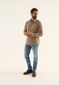 Chemise à carreaux boutonnée dans des tons marron et beige, associée à un jean bleu clair et des bottes en cuir marron, posant devant un fond neutre.