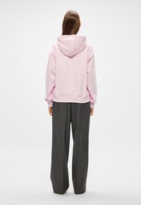 Ružová mikina s kapucňou vo voľnom strihu, skombinovaná s širokými šedými prúžkovanými nohavicami, ktoré vystihujú hladkú textúru látky a jednoduchý dizajn.