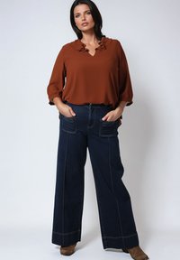Blouse couleur rouille avec un col froncé, associée à un jean à jambes larges bleu foncé doté de deux poches avant et de détails de couture contrastants.