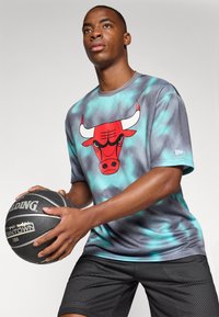 Grå og turkis tie-dye t-shirt med rød tyre-grafik, parret med sorte mesh shorts. Holder en sort basketball med hvide accenter.