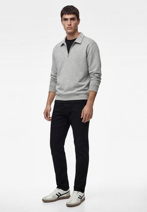 Młody mężczyzna stoi, ubrany w jasnoszary pulower z suwakiem do połowy, czarne spodnie oraz białe trampki z czarnymi paskami.