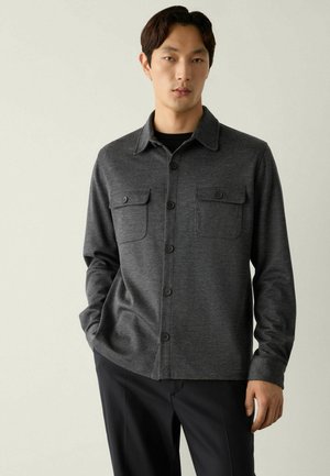 Mann trägt ein dunkelgraues Hemd mit Knöpfen und Brusttaschen sowie schwarze Hose, steht mit einer Hand in der Tasche vor einfarbigem Hintergrund.