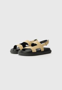 Sandales en suède beige avec semelle en caoutchouc noir, dotées de larges brides et d'une boucle ajustable à la cheville. Design plat avec un style à bout ouvert.