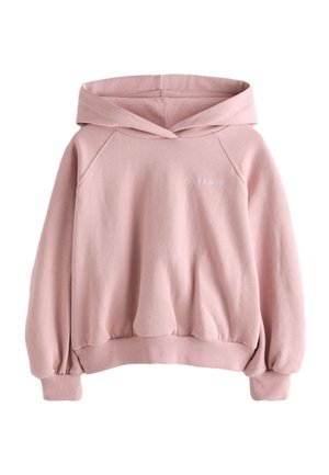 Pige med langt brunt hår iført en lyserød oversized hættetrøje med rynkede ærmer og gråvaskede jeans, står foran en grå baggrund.