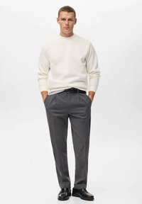 Suéter blanco de cuello redondo y pantalones grises de corte ajustado, complementados con un cinturón negro y zapatos negros, de pie contra un fondo sencillo.