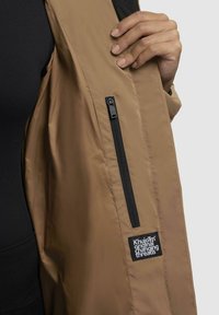 Brun jakke med mat finish, der har en sort lynlåslomme og en stoflabel. Inderforet viser en glat tekstilforing.