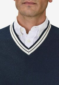 Marinblå väst med vit rand vid V-ringningen, lager över en vit skjorta med krage. Ribbad textur över hela plagget.