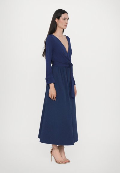 Navyblauwe wikkeljurk met een diepe V-hals, lange mouwen en een tailleband. Soepele stof; enkel-lengte rok. Gecombineerd met naakte hoge hakken sandalen.