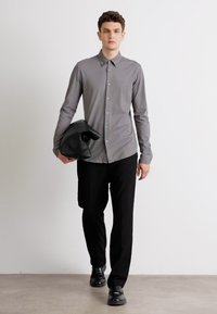 Grijze overhemd met kraag, lange mouwen en een gladde textuur, gecombineerd met zwarte broek en leren schoenen. Houdt een zwarte tas vast.