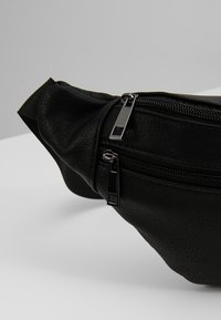 Sac banane en cuir synthétique noir avec deux compartiments zippés et des tirettes de fermeture éclair en métal argenté sur une surface gris blanc.