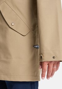 Un gros plan d'une main tenant le bord d'une veste beige, avec un bouton et une étiquette de marque près de l'ourlet.