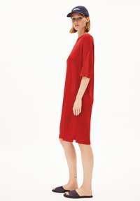 Robe tricotée rouge à manches courtes, longueur genou, avec des fentes sur les côtés à l'ourlet. Portée avec une casquette navy et des sandales noires à brides.