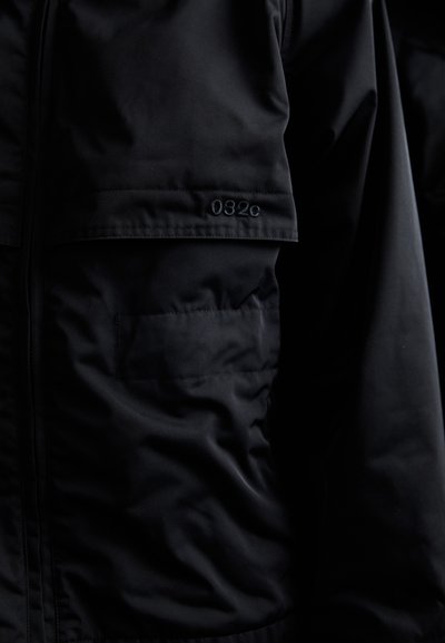 Sort jakke med en glat, mat finish, der har en brystlomme og broderet "032c" detalje nær skuldren.