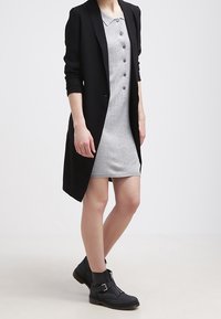Blazer lungo nero su un vestito grigio senza maniche con bottoni. Stivaletti neri alla caviglia con fibbie e finitura texturizzata su una suola piatta.