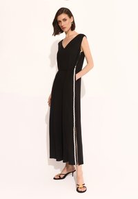 Czarny kombinezon z dekoltem w kształcie litery V, krótkimi rękawami, bocznymi kieszeniami oraz białym falbanym wykończeniem wzdłuż szwów bocznych; gładki materiał o luźnym kroju.