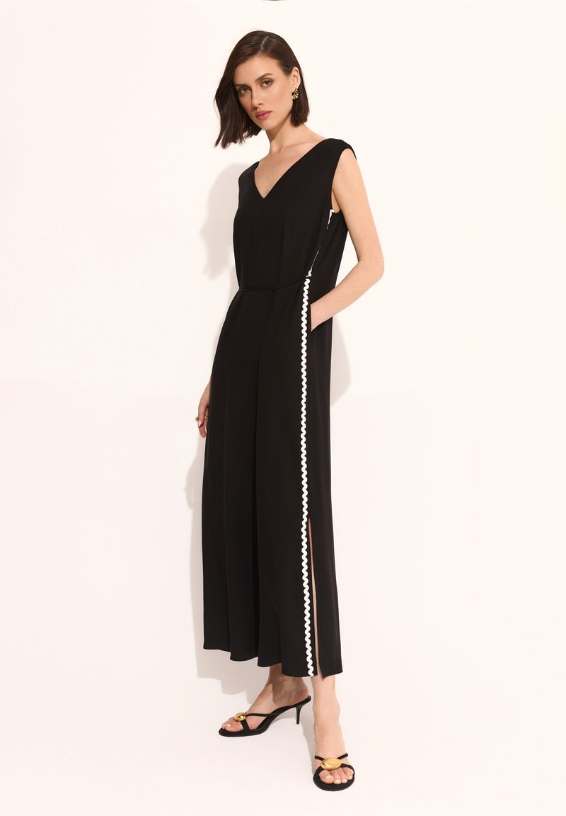 Czarny kombinezon z dekoltem w kształcie litery V, krótkimi rękawami, bocznymi kieszeniami oraz białym falbanym wykończeniem wzdłuż szwów bocznych; gładki materiał o luźnym kroju.