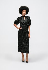 Crna haljina s sequinsom, s rubom do koljena, napučenim rukavima i visokim izrezom s mašnom. Nosena s crnim cipelama na špic.