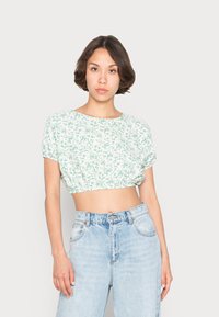 Háčkovaný crop top v bielej farbe s zeleným kvetinovým vzorom. Krátke rukávy a elastický lem. Spárovaný s vysokými modrými džínsami.
