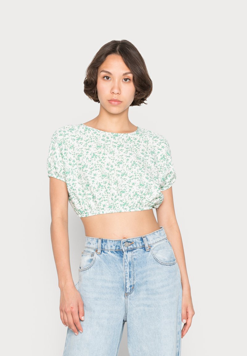 Háčkovaný crop top v bielej farbe s zeleným kvetinovým vzorom. Krátke rukávy a elastický lem. Spárovaný s vysokými modrými džínsami.