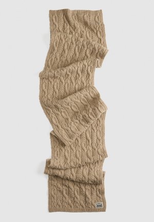 Gehaakte sjaal van beige wol, met een getextureerd kabelpatroon, afgewerkt met een geribbelde rand en een klein label in de onderste hoek.