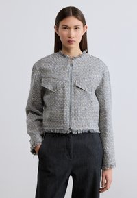 Κοντό γκρι σακάκι από καρό ύφασμα με φερμουάρ μπροστά, δύο τσέπες στο στήθος, ακατέργαστες λεπτομέρειες στο τελείωμα και υφή με υφή. Συνδυάζεται με σκούρο παντελόνι.