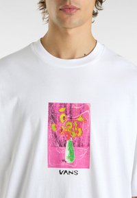Vit t-shirt med en rosa grafik som föreställer en vas med blommor, gröna detaljer och ordet "VANS" tryckt nedanför. Kort ärm och rundad hals.