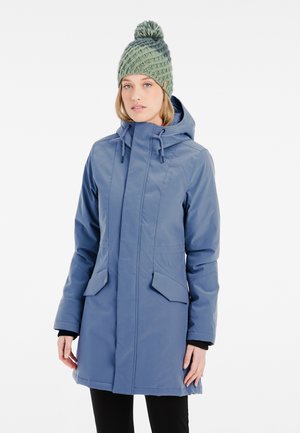 Blaue wasserdichte Jacke mit einer Kapuze, Reißverschluss, zwei vorderen Taschen und verstellbaren Kordeln. Kombiniert mit einer grünen Strickmütze.