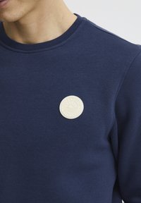 Tamsiai mėlynas džemperis su apvaliu, tiksliai tekstūruotu baltu logotipu ant krūtinės. Pagamintas iš lygaus audinio su klasikinėmis apykaklėmis.