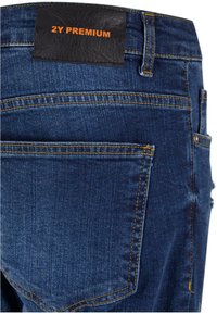 Vaqueros de denim azul oscuro con etiqueta de la marca en naranja en la cintura, costuras visibles y dos bolsillos traseros. Ligeramente desgastados cerca del dobladillo.