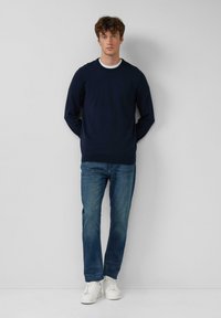 Giovane uomo in piedi contro una parete bianca, indossa un maglione blu navy, jeans blu e sneaker bianche, con le mani dietro la schiena.