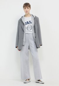 Blazer grigio oversize sopra una felpa grigia con stampa "ZAL University", abbinato a pantaloni sportivi grigi larghi e ballerine argentate.