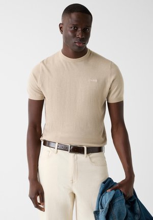 Man met beige shirt met korte mouwen en crèmekleurige broek met een bruine riem, houdt een blauwe spijkerjack in zijn rechterhand tegen een witte achtergrond.