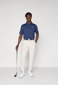 Granatowa koszulka polo z kołnierzykiem i subtelną teksturą, połączona z beżowymi spodniami, białymi sneakersami i trzymana kije golfowego.