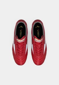 Pantofi de fotbal roșii cu un brâu din piele netedă, accente albe pe laterale și o talpă texturată. Prezintă un sistem de șireturi roșii și un logo auriu.