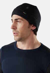 Ribbed black knitted beanie with a small logo on the side and colorful accent stripes; worn by a man with short hair and a blue shirt.